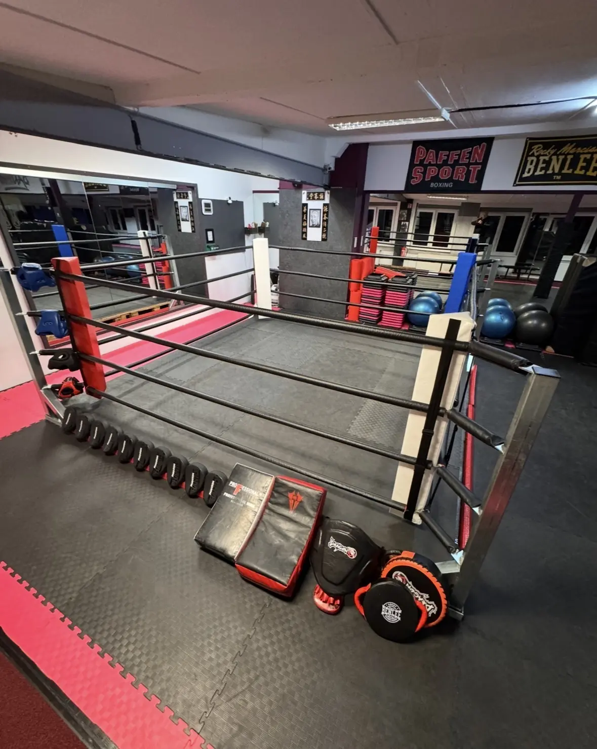 Boxring mit roten und weißen Ecken, umgeben von Trainingsgeräten und Matten in einem Fitnessstudio.