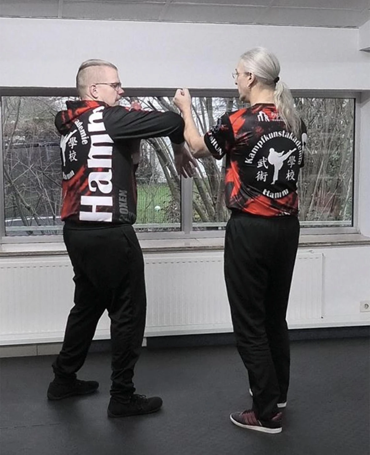 Zwei Personen in Trainingsanzügen, die Wing Tsun Techniken in einem Übungsraum demonstrieren, mit Fokus auf die Armbewegungen.