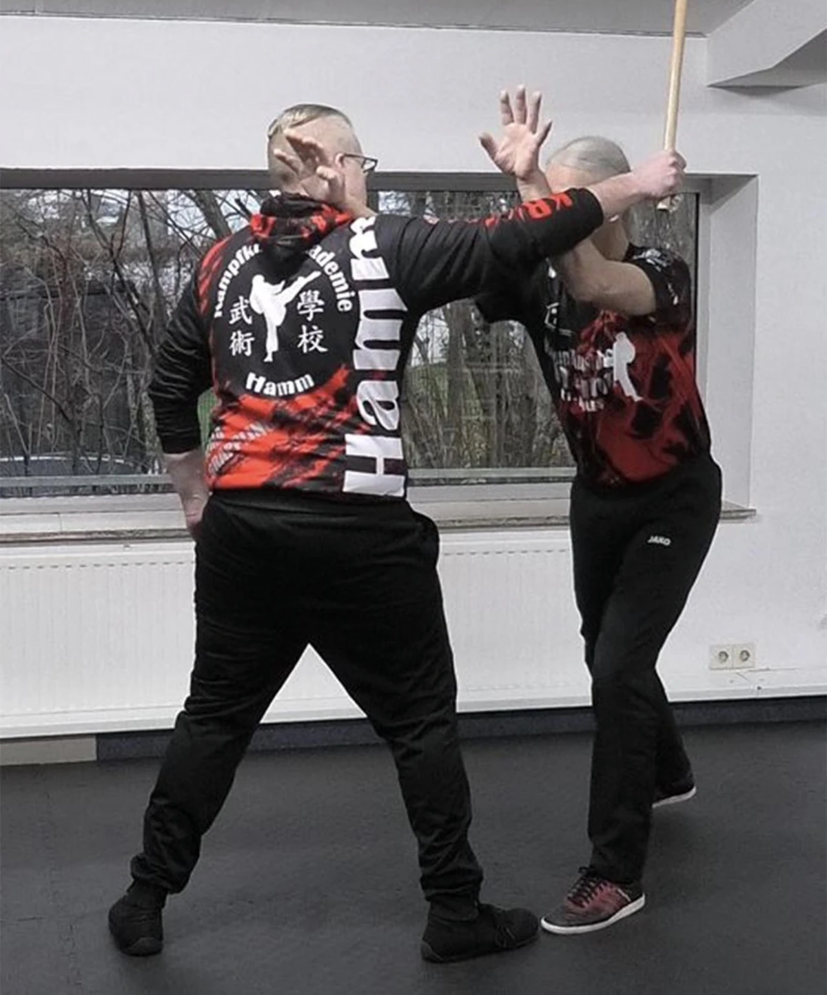 Zwei Personen in Kampfkunstkleidung üben Wing Tsun Techniken mit einem Stock in einem Trainingsraum.
