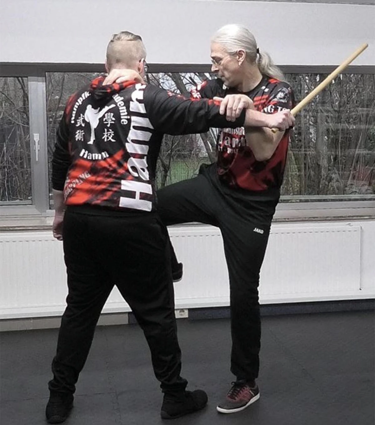Zwei Personen in Kampfkunstkleidung üben Wing Tsun Techniken mit einem Stock in einem Trainingsraum.
