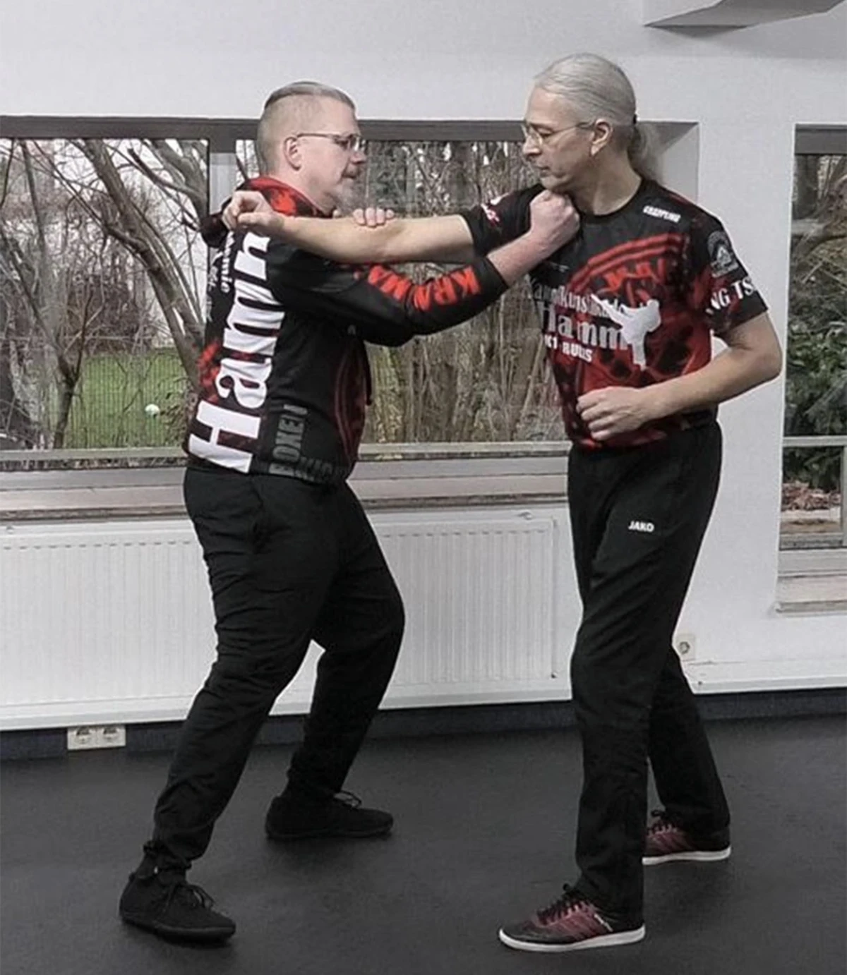 Zwei Personen in Kampfkunstkleidung üben Wing Tsun Techniken in einem Trainingsraum. Beide tragen sportliche Oberteile mit dem Schriftzug 'Hamm'.