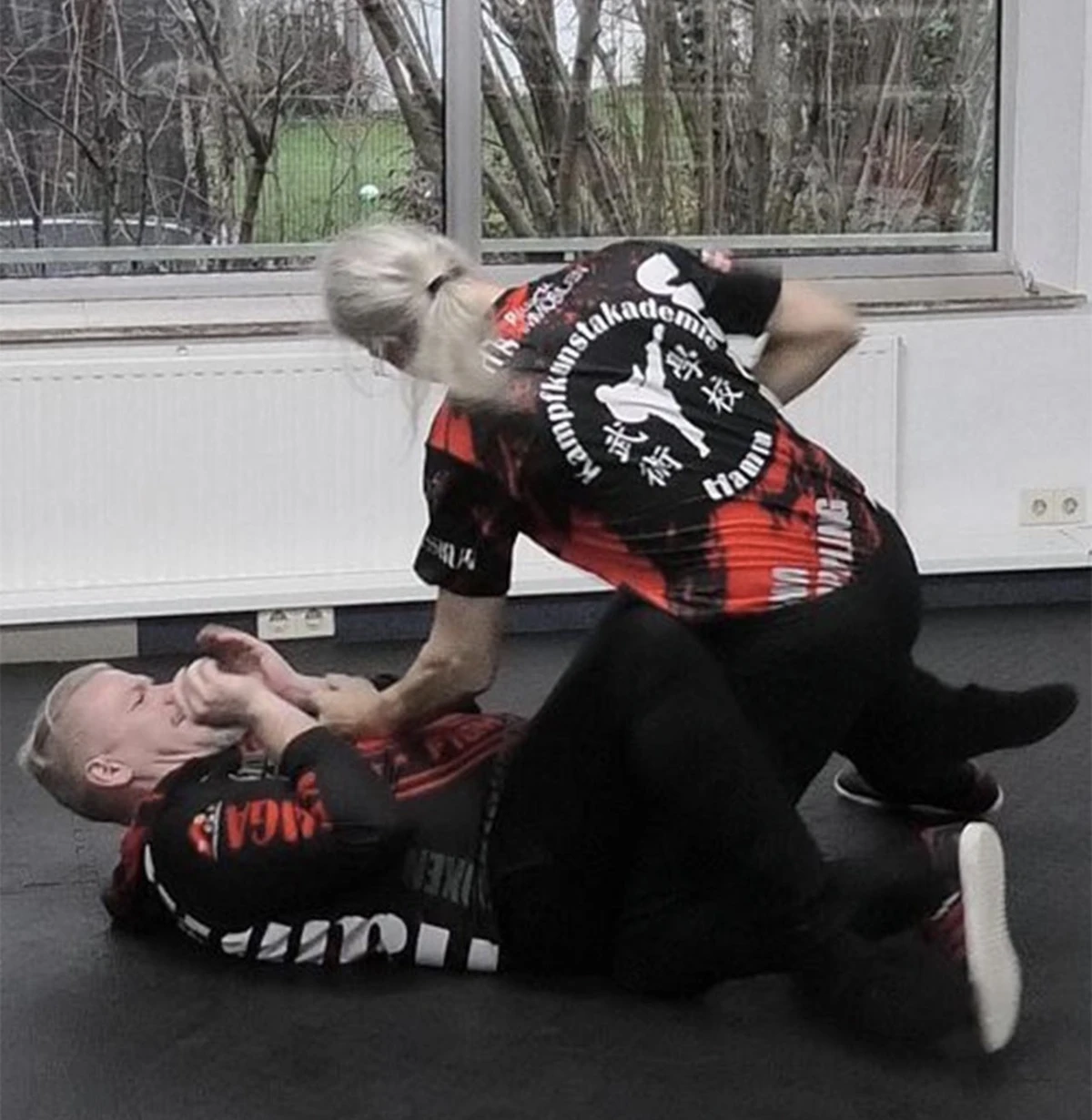 Zwei Personen in Kampfsportkleidung, die eine Technik im Wing Tsun demonstrieren, während sie auf einer Matte trainieren.