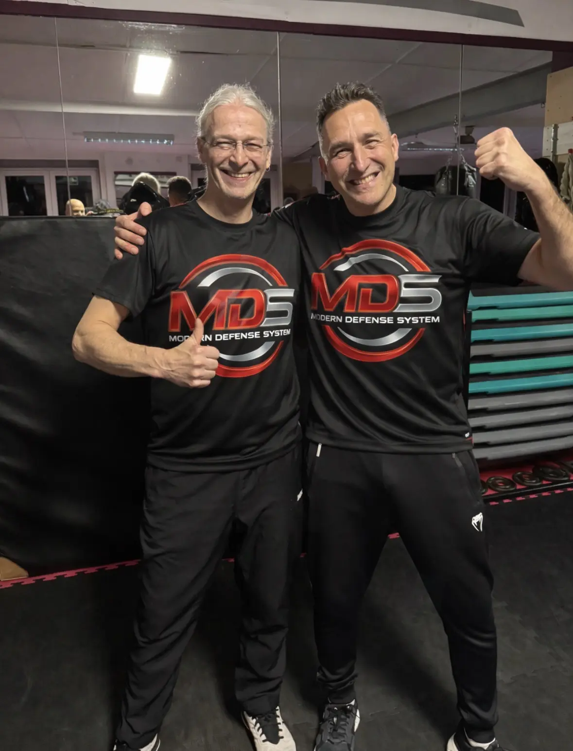 Zwei Männer in schwarzen T-Shirts mit dem Logo 'MDS' stehen nebeneinander und zeigen Daumen hoch, während sie lächeln.